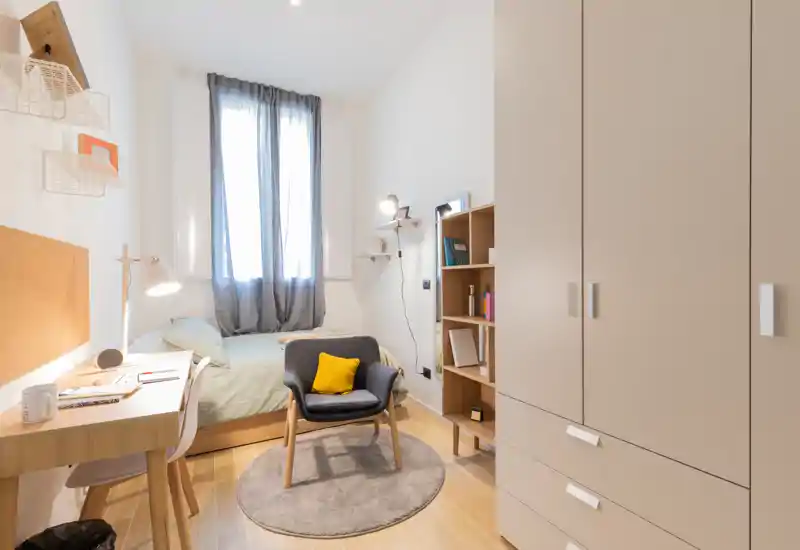 Dormitorio pequeño y luminoso con cama individual, escritorio, armario y un cómodo sillón con cojín amarillo. La luz natural a través de las cortinas translúcidas crea un ambiente cálido e ideal para un estudiante o una persona sola.