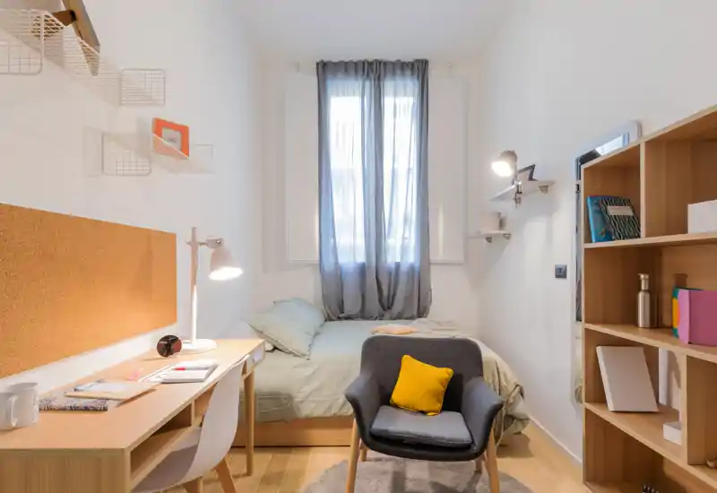 Dormitorio individual luminoso y bien decorado con cama cómoda bajo una ventana con cortina fina, escritorio, estantería y un sillón acogedor con cojín amarillo — ideal para estudiante o ocupante único.