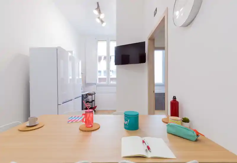 Bright, modern kitchen/dining corner with a wooden table set for casual use, refrigerator and window in the background, and a wall-mounted TV—ideal for small-apartment listings.