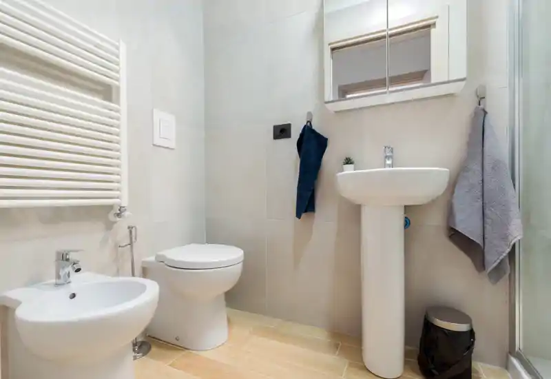 Clean, modern bathroom with a pedestal sink, toilet and bidet, neutral tiles and a mirrored cabinet above the sink.