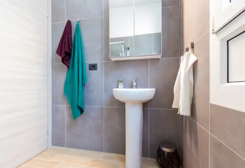 Compact, modern bathroom with a pedestal sink, mirrored cabinet and neutral grey tiles; well-lit and neatly styled for rental photos.