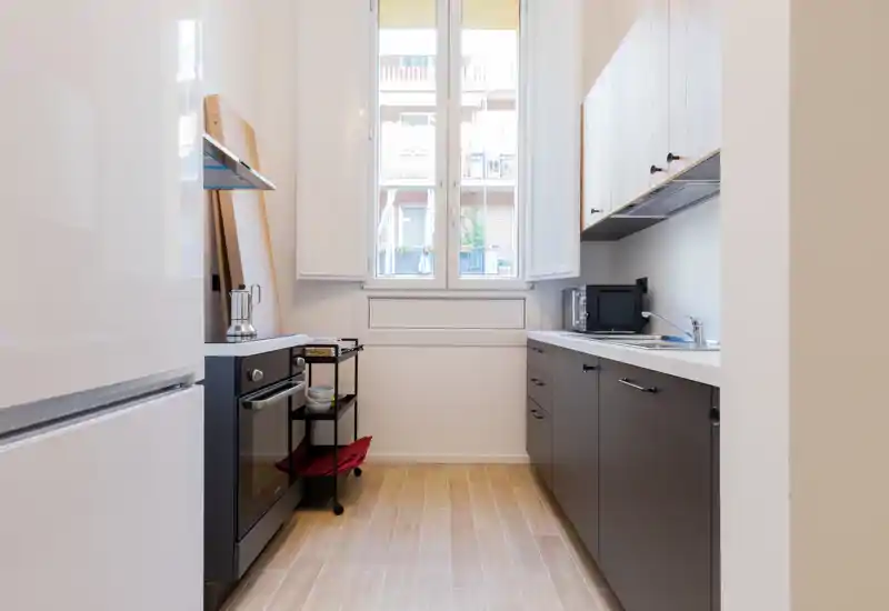 Bright, modern galley kitchen with dark lower cabinets, light wood-look floor and a window providing natural light. Compact layout includes oven, microwave, fridge and a small rolling cart — ideal for a functional city apartment.