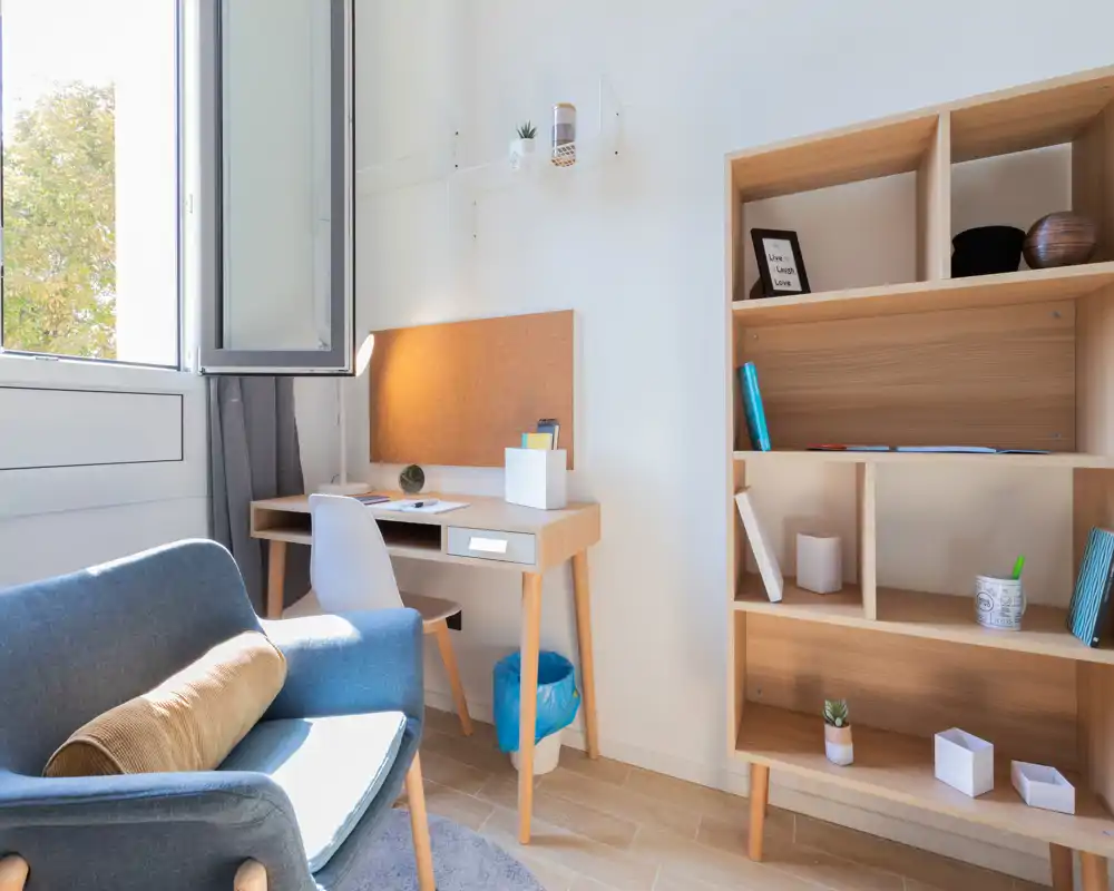 Bright, modern living area with a comfortable armchair, compact desk by an open window and a wooden shelving unit—ideal for a cozy work-and-relax corner.
