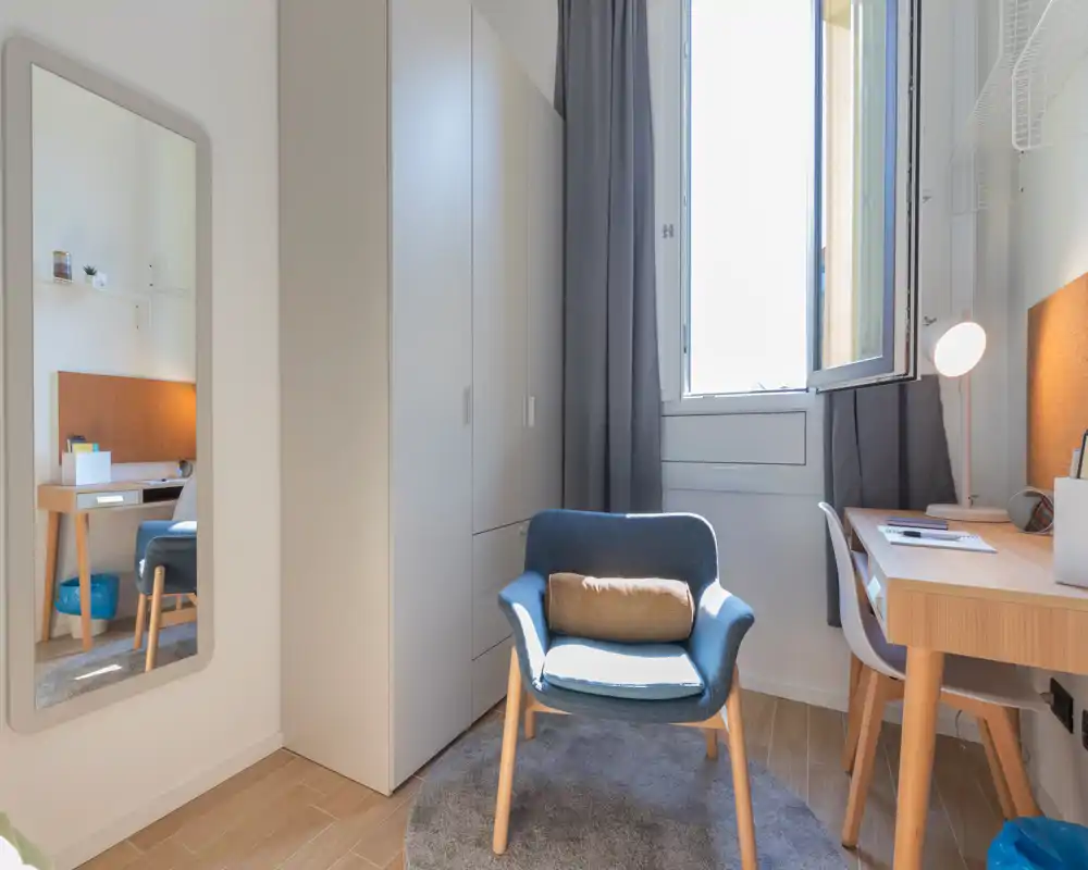 Small, well-lit bedroom corner featuring a comfortable armchair, desk with lamp and a wardrobe; the bed is visible only as a reflection in the wall mirror. Fresh, minimal décor with natural light from the window.
