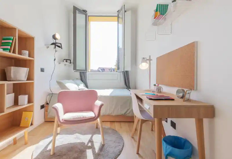 Camera da letto luminosa e accogliente con letto singolo sotto una finestra aperta, scrivania in legno con lampada e una comoda poltroncina rosa su un tappeto rotondo. Arredamento in stile scandinavo, scaffalature in legno e atmosfera ariosa, ideale per uno studente o un occupante singolo.