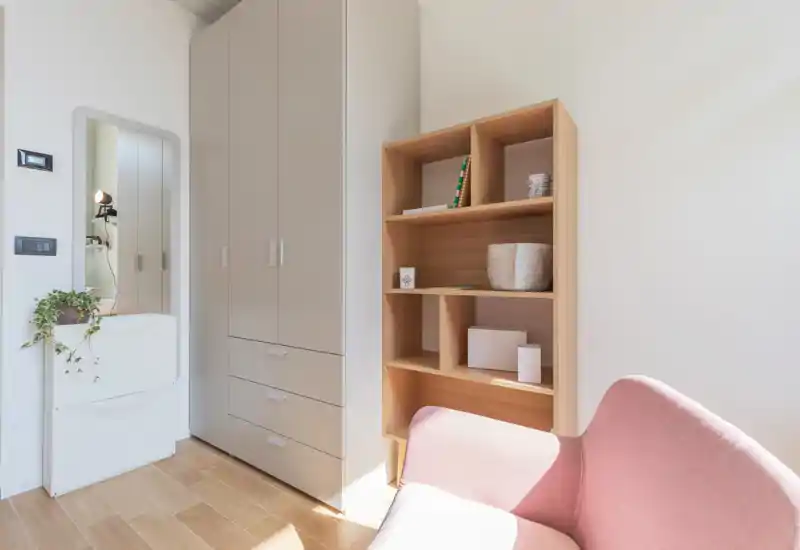 Angolo living moderno e luminoso con una poltrona rosa, mensole aperte e un armadio alto; linee pulite e luce naturale creano un ambiente accogliente.