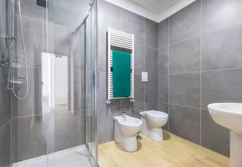 Modern bathroom with a glass shower enclosure, wall-mounted towel radiator with a green towel, bidet, toilet and pedestal sink; light wood-look floor tiles and large grey wall tiles create a clean, contemporary look.