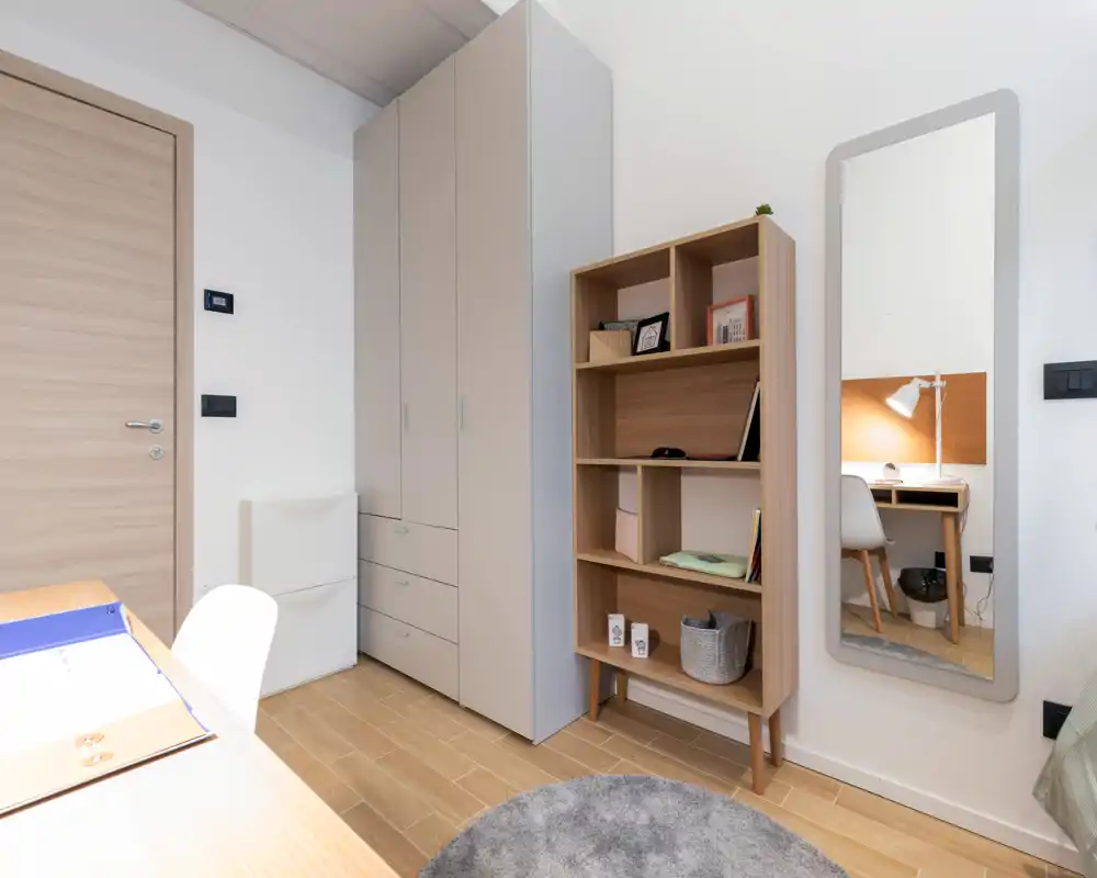 Coin de chambre lumineux et moderne avec une grande armoire, une étagère ouverte et un miroir mural reflétant un petit espace de travail ; tons neutres et sol en bois pour une ambiance apaisante.