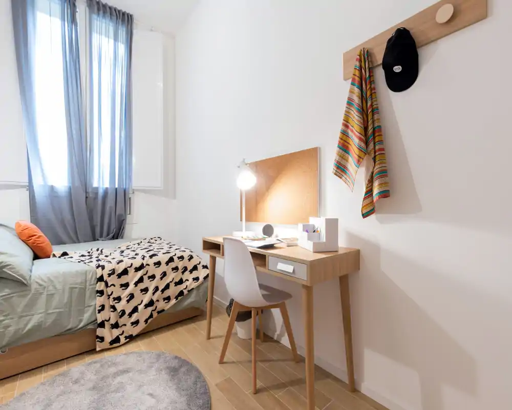 Chambre simple moderne et lumineuse avec lit, bureau et lumière naturelle. Lignes épurées, sol en bois et textiles confortables, parfaite pour un étudiant ou un professionnel.