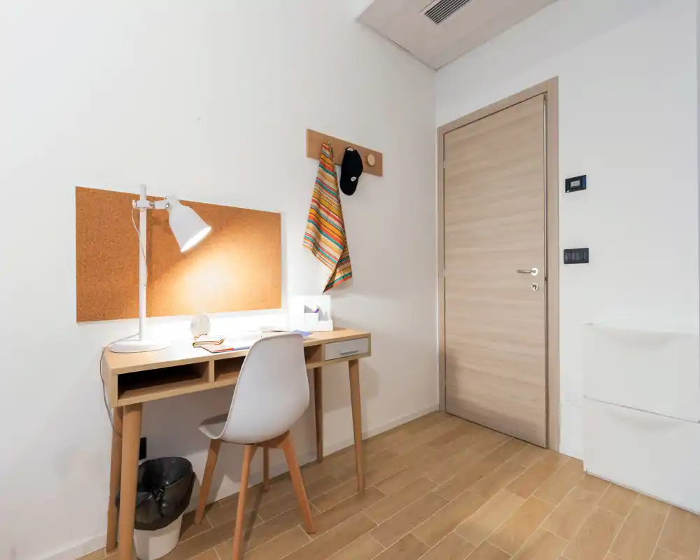 Coin de chambre compact avec bureau en bois, chaise, panneau en liège et lampe ; espace de travail lumineux et soigné près de la porte.