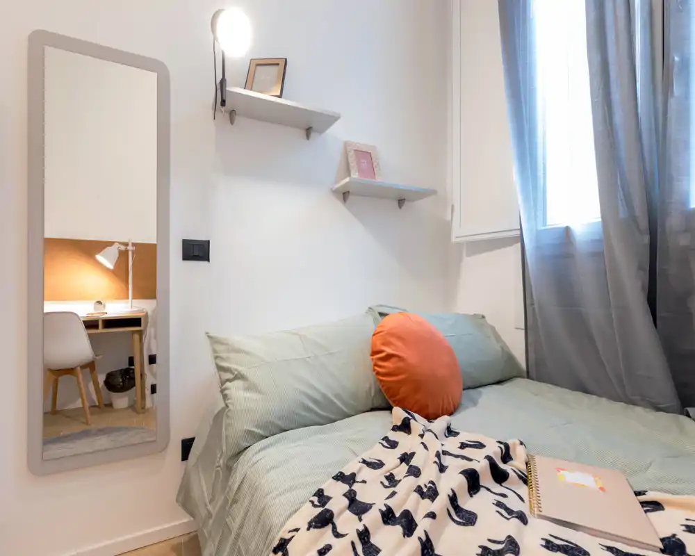 Chambre lumineuse et soignée avec un lit double bien fait, coussin rond décoratif et plaid à motif. Une glace pleine longueur reflète un coin bureau et la lumière naturelle entre par la fenêtre.
