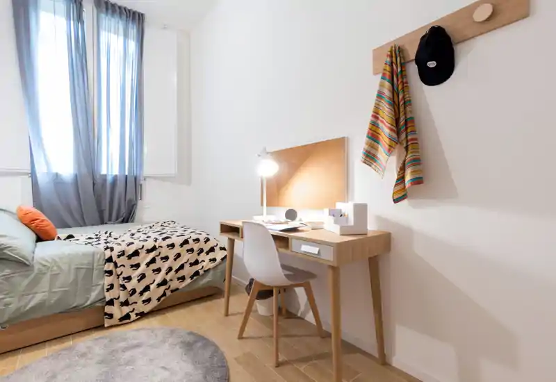 Chambre simple moderne et lumineuse avec lit, bureau et lumière naturelle. Lignes épurées, sol en bois et textiles confortables, parfaite pour un étudiant ou un professionnel.