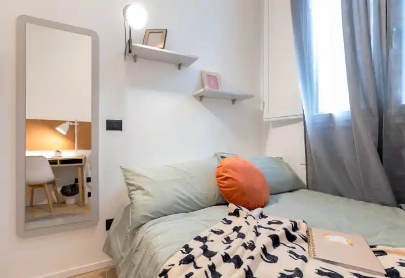 Chambre lumineuse et soignée avec un lit double bien fait, coussin rond décoratif et plaid à motif. Une glace pleine longueur reflète un coin bureau et la lumière naturelle entre par la fenêtre.