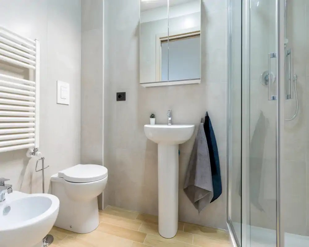 Salle de bains moderne et bien éclairée avec lavabo sur pied, toilette, bidet et cabine de douche en verre. Carrelage neutre et sol imitation bois pour un rendu propre et contemporain.
