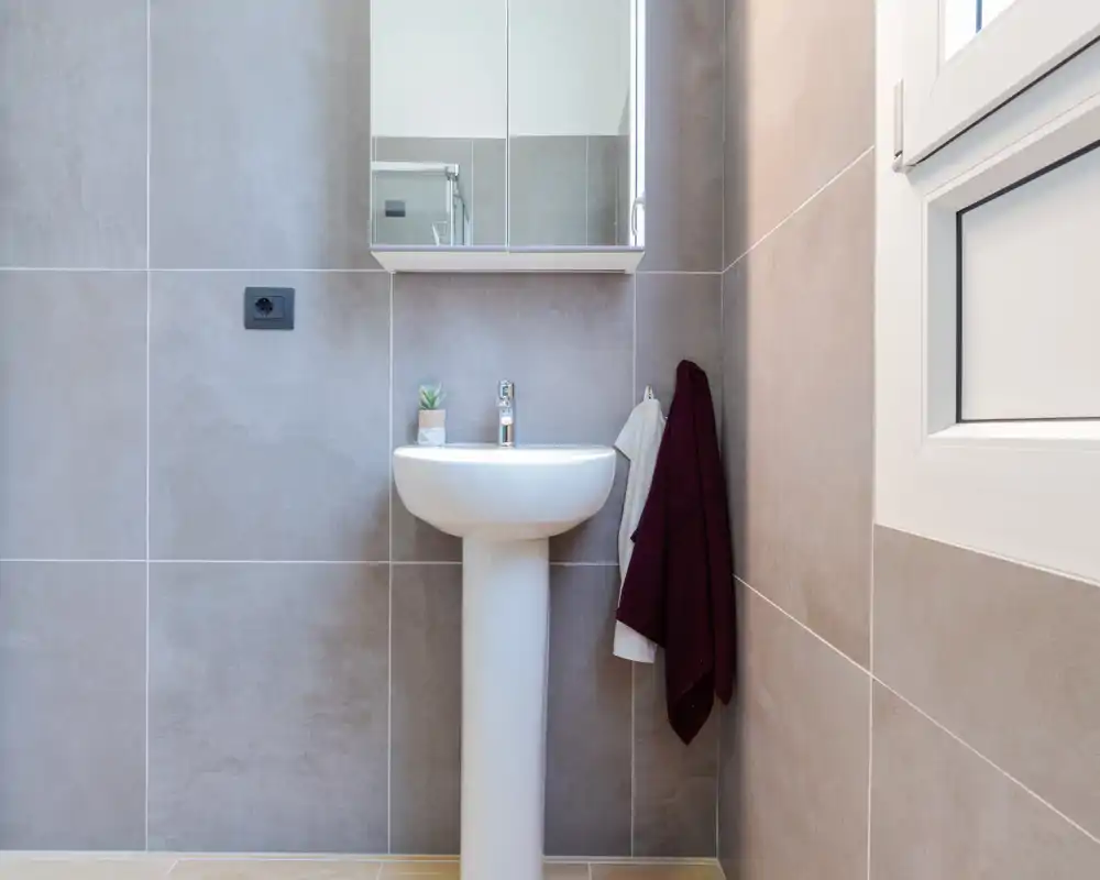 Salle de bain moderne et lumineuse avec lavabo sur colonne, armoire miroir et grandes dalles neutres ; des serviettes sobres apportent une touche chaleureuse.