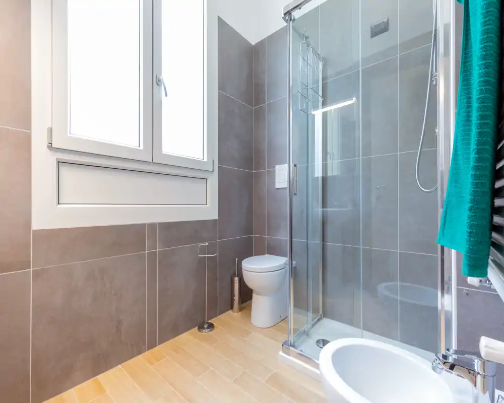 Salle de bain moderne et lumineuse avec cabine de douche vitrée, WC suspendu, bidet et grande fenêtre dépoli ; sol effet bois et carrelage mural gris pour un style contemporain.