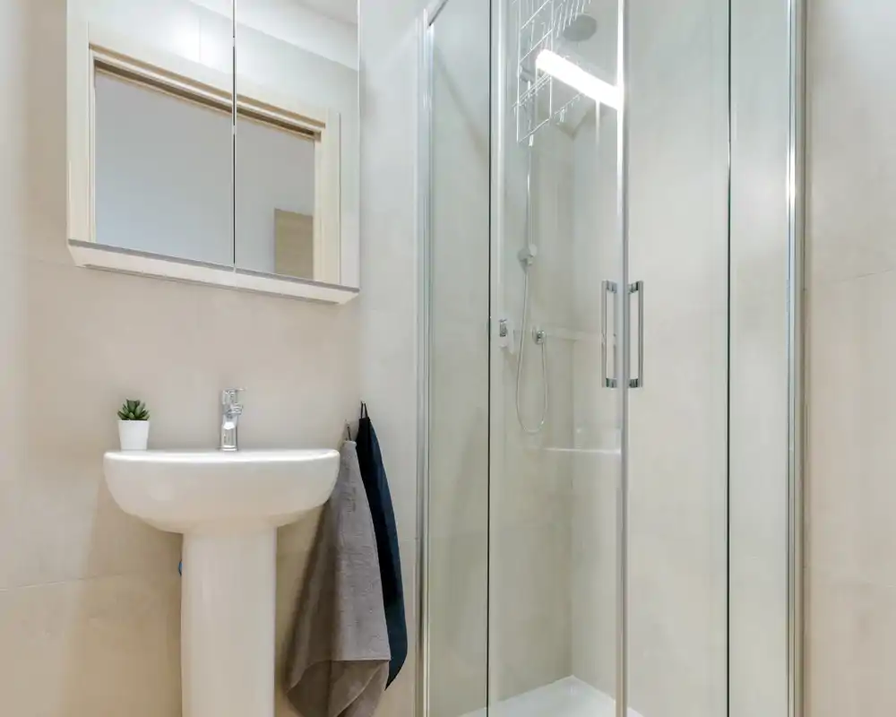 Salle de bains moderne et lumineuse avec lavabo sur colonne, armoire-miroir et douche à cabine vitrée ; carrelage neutre et style soigné.
