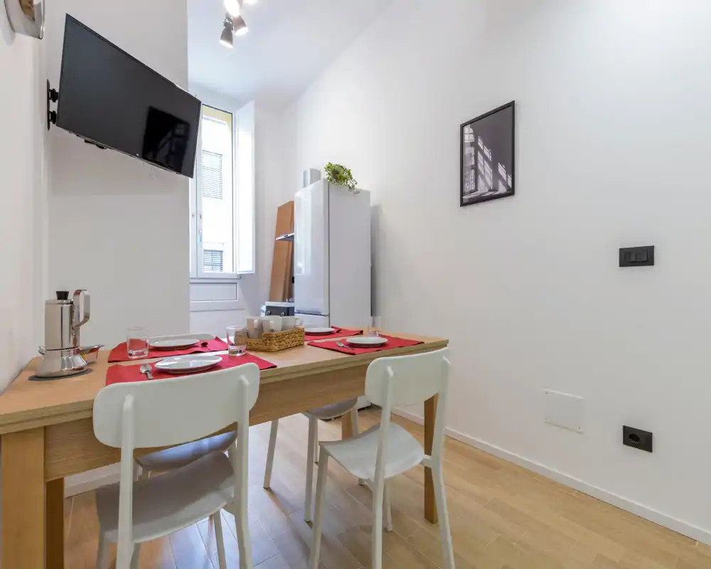 Coin repas compact avec table en bois dressée pour quatre, chaises blanches modernes et fenêtre lumineuse ; éléments de kitchenette en arrière-plan, parfait pour un appartement urbain.