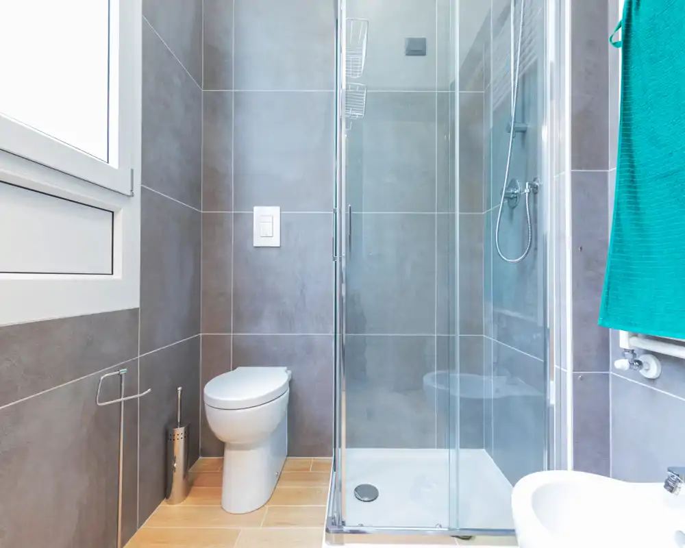 Salle de bains moderne et compacte avec douche vitrée, toilette murale, bord de bidet visible et une fenêtre lumineuse apportant de la lumière naturelle. Carrelage neutre et sol imitation bois pour une ambiance contemporaine.