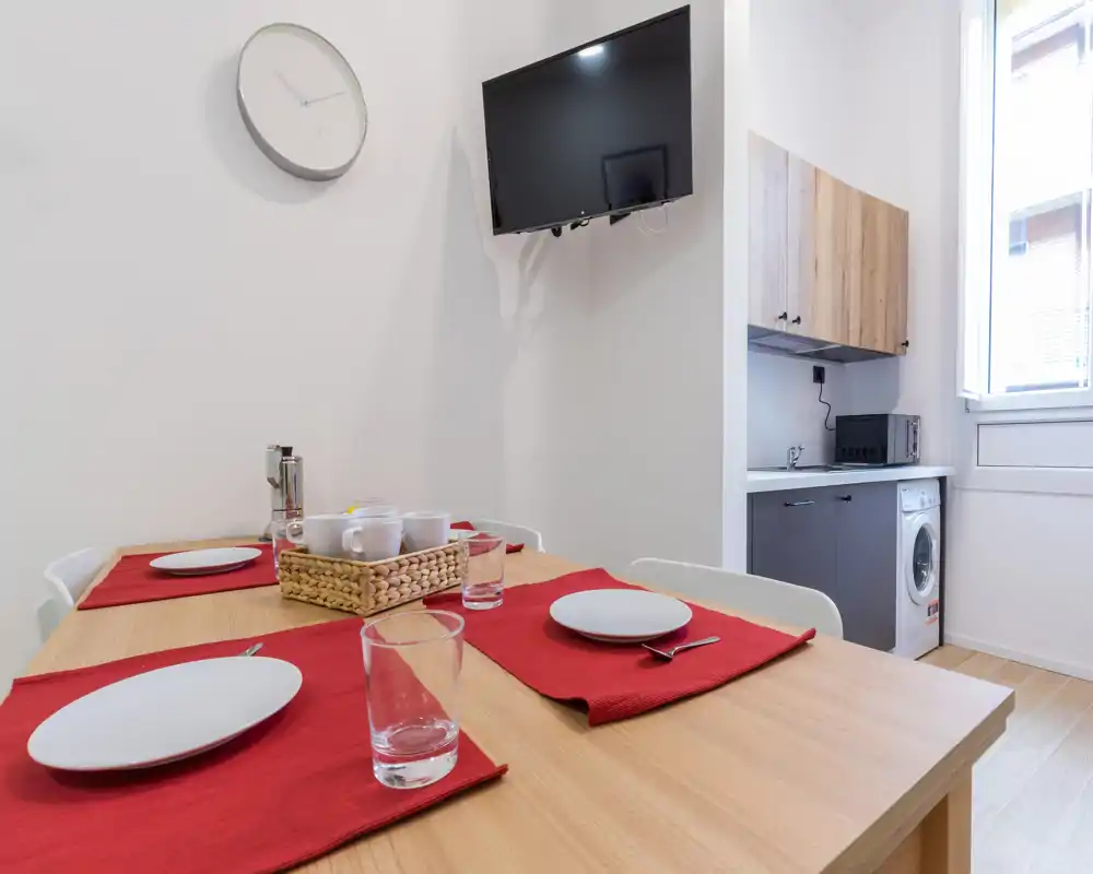 Coin repas lumineux et moderne avec une table en bois pour quatre, des sets de table rouges et une kitchenette compacte en arrière-plan.