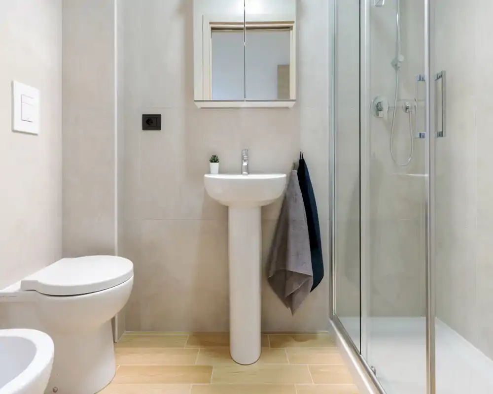 Salle de bains moderne et lumineuse avec lavabo sur colonne, miroir-armoire, WC et douche avec paroi vitrée. Carrelage neutre et sol effet bois pour un rendu propre et contemporain.