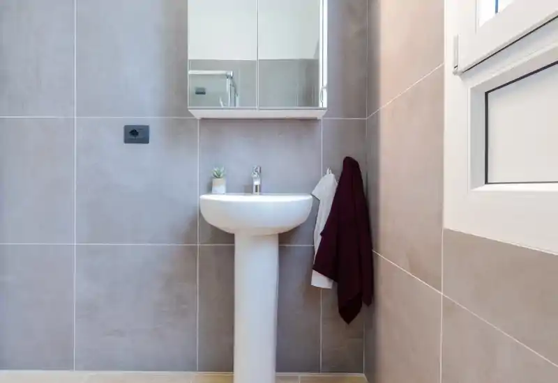 Bright, modern bathroom with a pedestal sink, mirrored cabinet and neutral large-format tiles; simple towels add a touch of warmth.