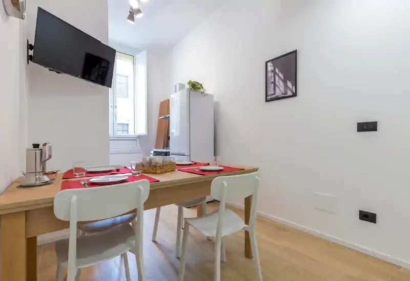 Compact dining area with a wooden table set for four, modern white chairs and a bright window; small kitchenette elements visible in the background make it ideal for a city apartment.