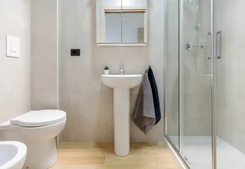 Bright, modern bathroom with a pedestal sink, wall-mounted cabinet mirror, toilet and a glass-enclosed shower. Neutral tiles and wood-effect floor create a clean, contemporary look.