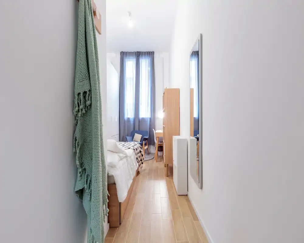 Dormitorio individual estrecho y luminoso con cama bien hecha, sillón junto a la ventana y pequeño escritorio. Mobiliario de estilo nórdico y luz natural crean un ambiente acogedor, ideal para alquileres de corta estancia.