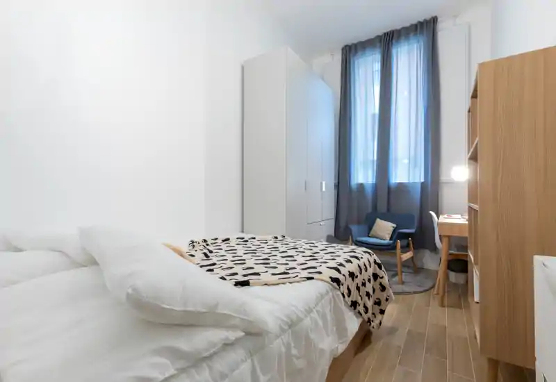 Dormitorio luminoso y bien presentado con cama doble confortable, manta de estampado, armario y un sillón acogedor junto a la ventana.