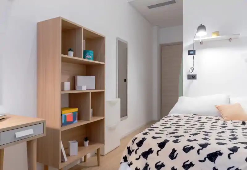 Dormitorio luminoso y compacto con manta estampada y lámpara de pared; estantería de madera clara para almacenaje, el espacio se siente limpio y acogedor.