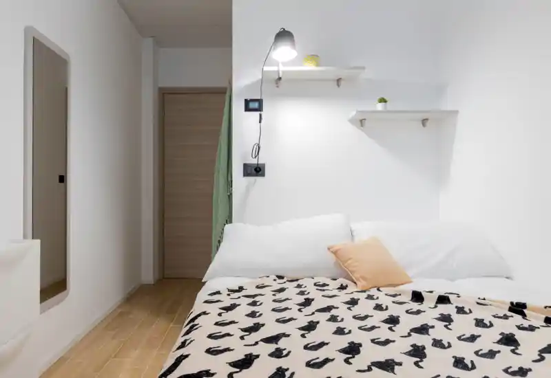 Dormitorio luminoso y minimalista con una gran cama vestida con ropa blanca, una manta estampada y un cojín cálido; estanterías sencillas y una lámpara de lectura completan el ambiente acogedor.
