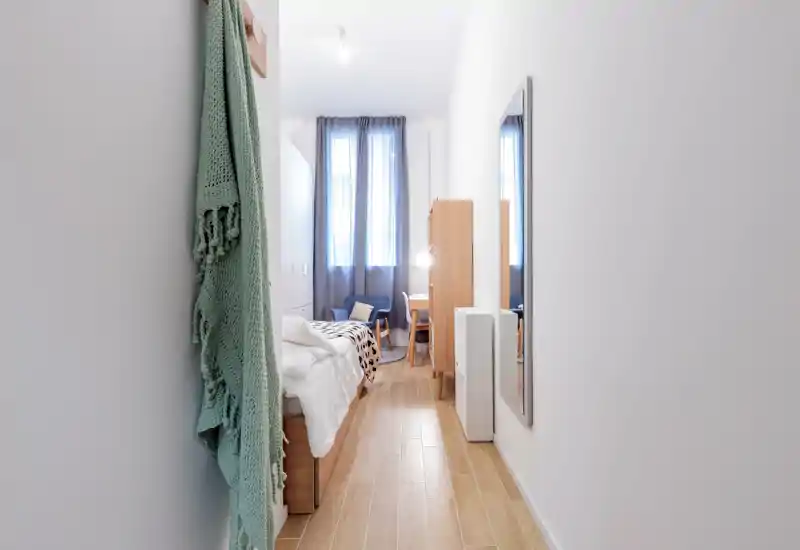 Dormitorio individual estrecho y luminoso con cama bien hecha, sillón junto a la ventana y pequeño escritorio. Mobiliario de estilo nórdico y luz natural crean un ambiente acogedor, ideal para alquileres de corta estancia.