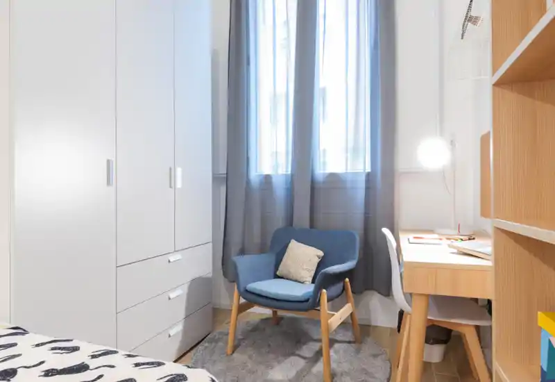 Rincón de dormitorio compacto y bien decorado: parte de la cama visible en el borde, butaca cómoda sobre alfombra redonda, escritorio de madera con lámpara y estanterías — luminoso, moderno y acogedor.