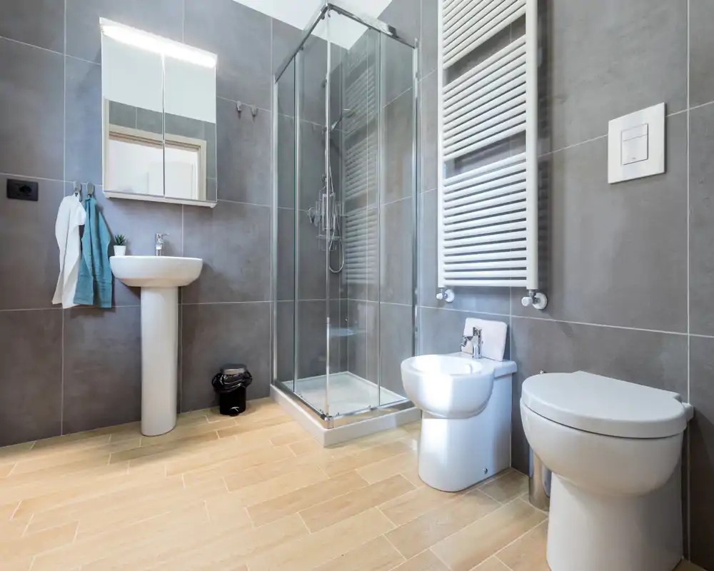 Bagno moderno e ben illuminato con box doccia in vetro, lavabo a colonna, bidet e WC su piastrelle grandi grigie e pavimento effetto legno chiaro.
