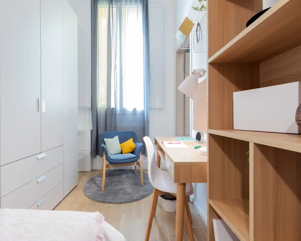 Angolo camera da letto luminoso e moderno con letto parzialmente visibile, scrivania e scaffalature in legno e una poltroncina accogliente vicino alla finestra, ideale per uno studente o single.