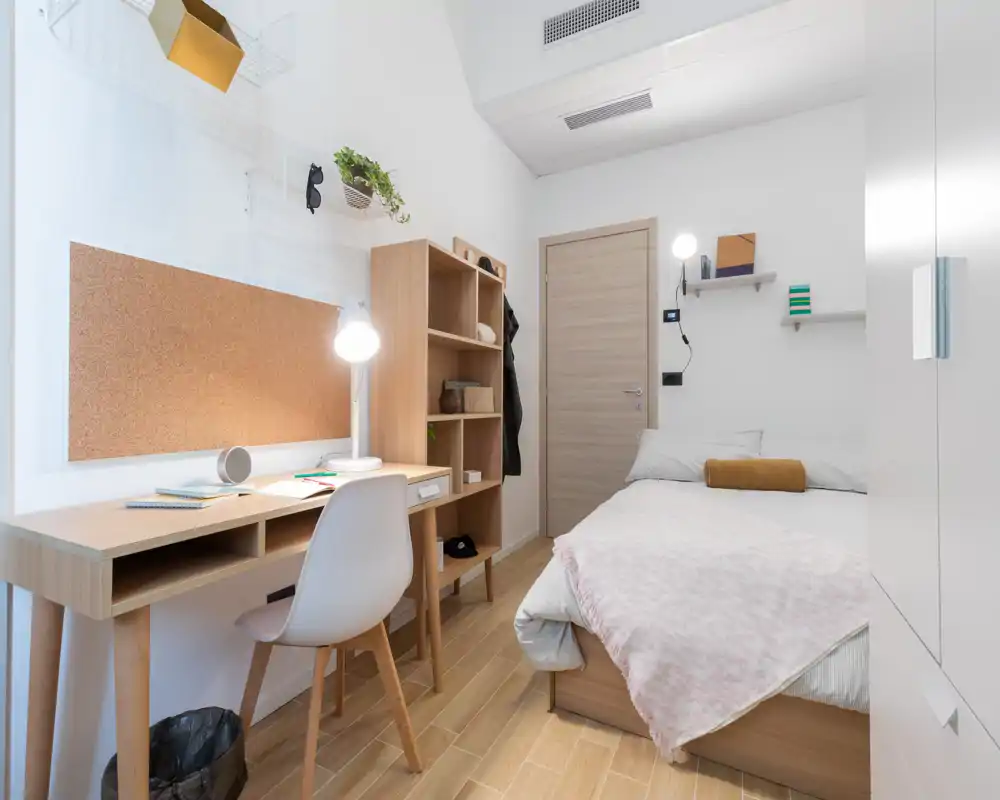 Camera da letto luminosa e compatta con letto singolo, mobili in legno e una scrivania ordinata — ideale per uno studente o una persona sola. Tessuti morbidi, mensole e uno stile minimalista che massimizza lo spazio.