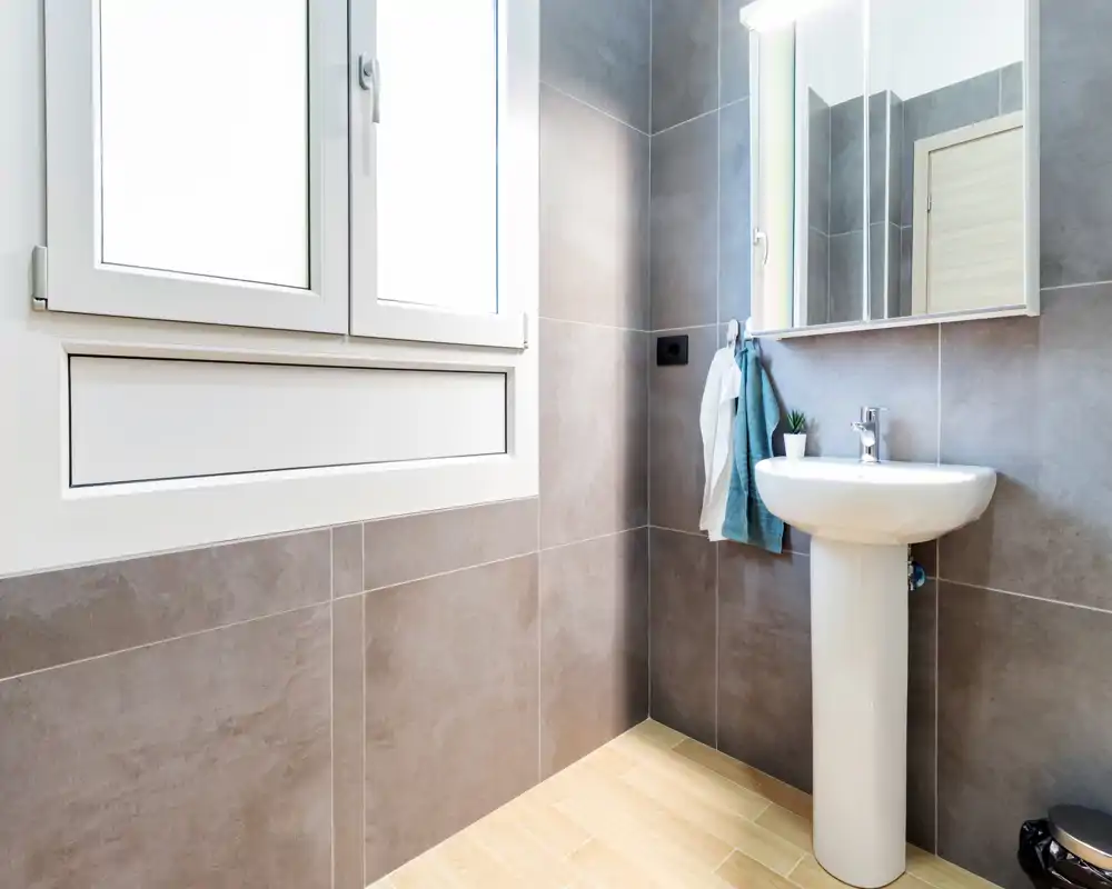 Bagno moderno e luminoso con lavabo a colonna, grande specchio e piastrelle grigie neutre. La luce naturale entra da una finestra con cornice bianca, creando un ambiente pulito e arioso.