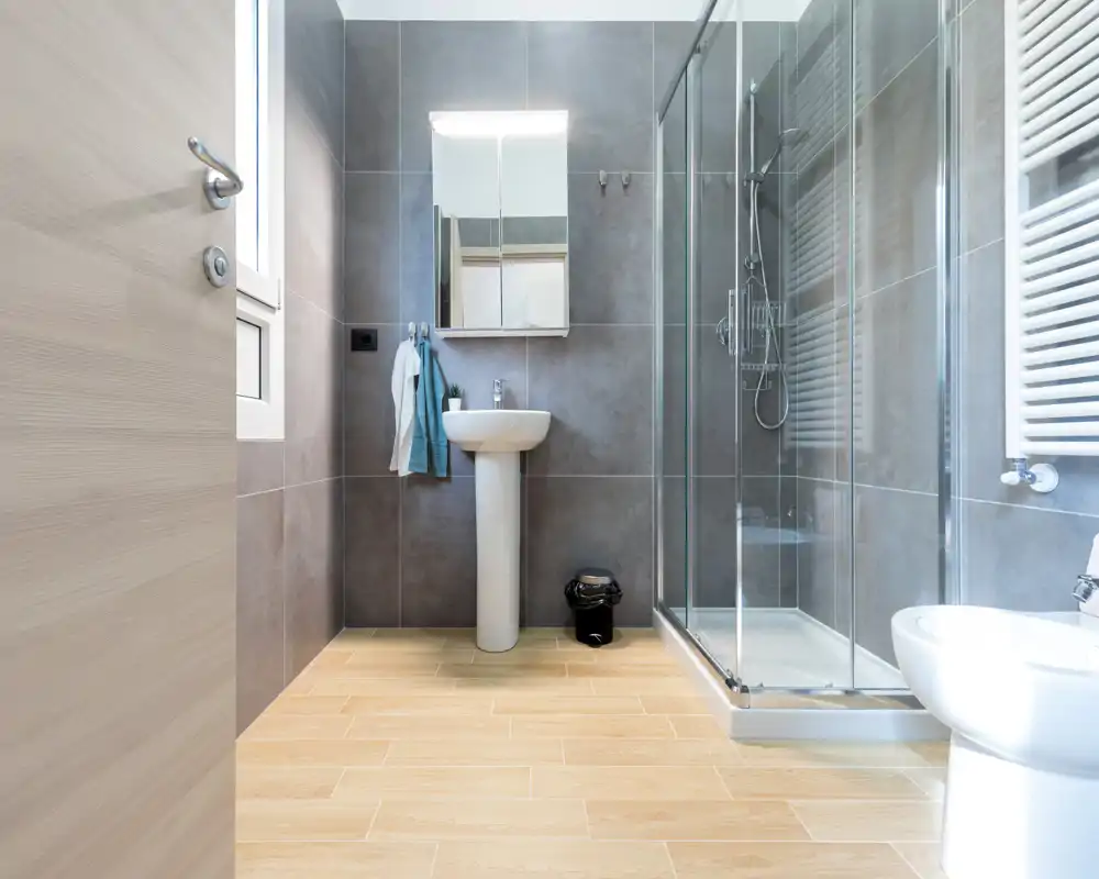 Bagno moderno e ben illuminato con cabina doccia in vetro, lavabo a colonna e piastrelle nei toni neutri; pavimento effetto legno che dona calore.