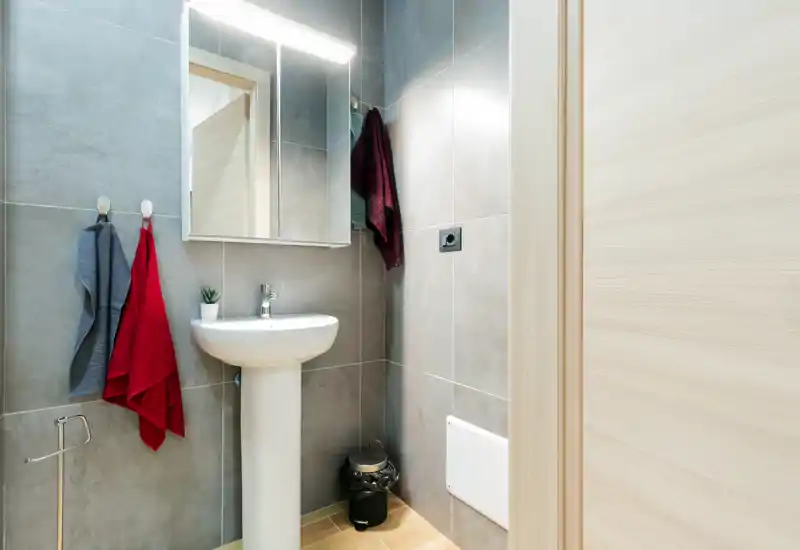 Compact modern bathroom with a pedestal sink, illuminated mirror, tiled walls and hanging towels; bright, clean and functional.