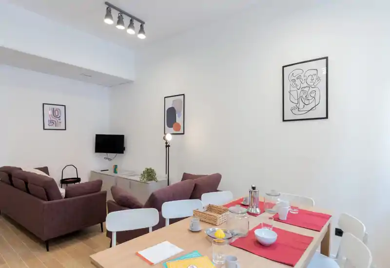 Bright, modern living area with a comfortable two-seat sofa, armchair and a dining table in the foreground. Neutral walls, framed prints and soft lighting give the space a calm, welcoming feel ideal for rental photos.