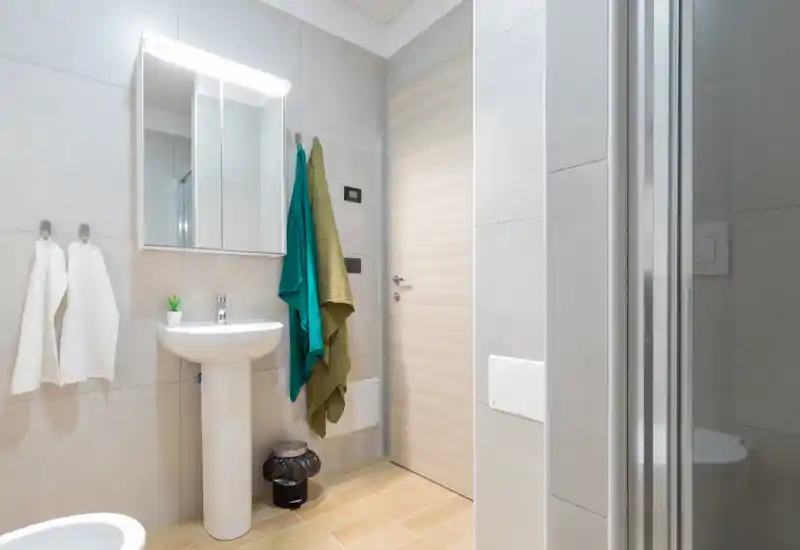 Compact modern bathroom with a pedestal sink, illuminated mirror cabinet, wall-mounted towels and a glass-enclosed shower partially visible at right; light neutral tiles and wood-effect floor create a clean, contemporary look.