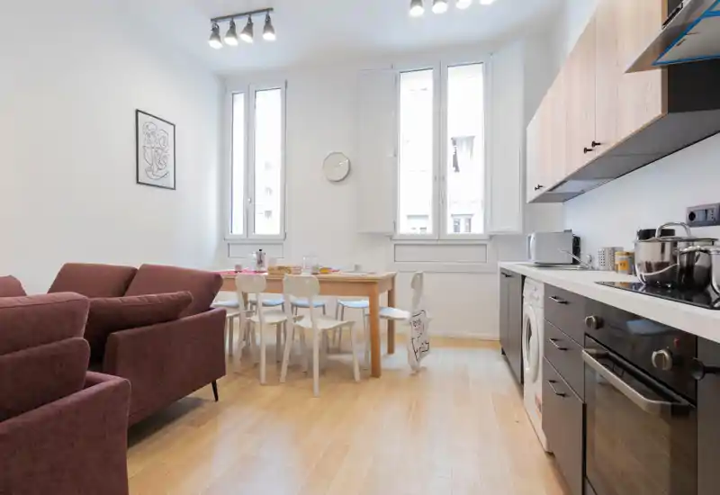 Bright open living area with a compact modern kitchen and a dining table; comfortable plum sofas and natural light create a welcoming space ideal for short-term rentals.