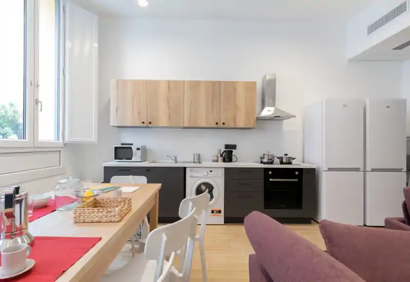 Modern open kitchen with wooden cabinets, built-in appliances and a dining table by the window; bright, clean and tastefully furnished for short stays.