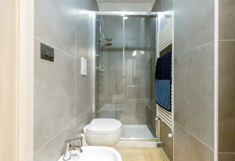 Compact modern bathroom with a glass-enclosed shower, wall-mounted toilet and bidet, neutral gray tiles and a heated towel rail with towels.