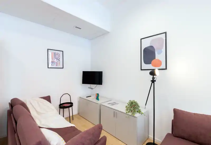 Compact, well-styled living room with a plum sofa, wall-mounted TV, and a floor lamp. Neutral walls, modern prints and a low cabinet create a tidy, welcoming lounge area ideal for relaxing or watching TV.