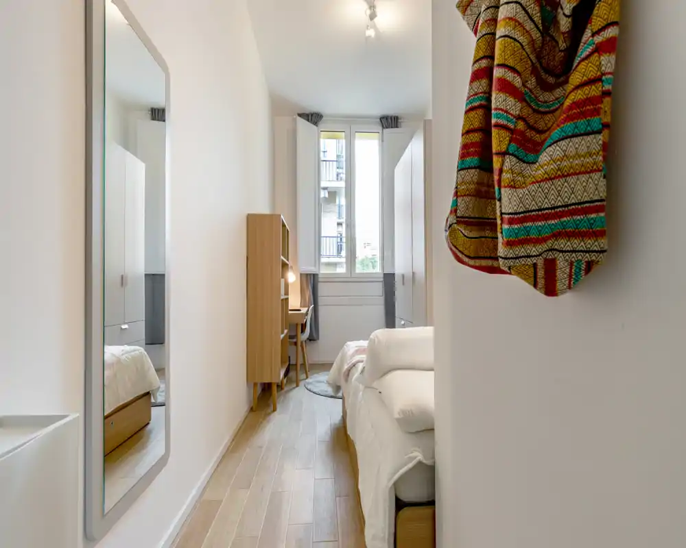 Dormitorio moderno y luminoso con cama individual a la derecha, escritorio de madera y armario junto a la ventana, y un gran espejo a la izquierda — limpio, espacioso y bien iluminado.