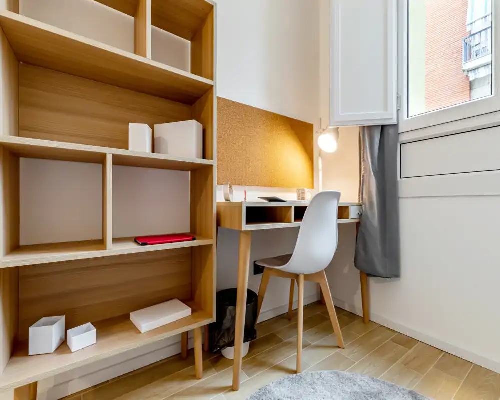 Rincón de oficina en casa luminoso y minimalista con escritorio de madera, corcho para notas y estanterías abiertas junto a una gran ventana — ideal para concentrarse.