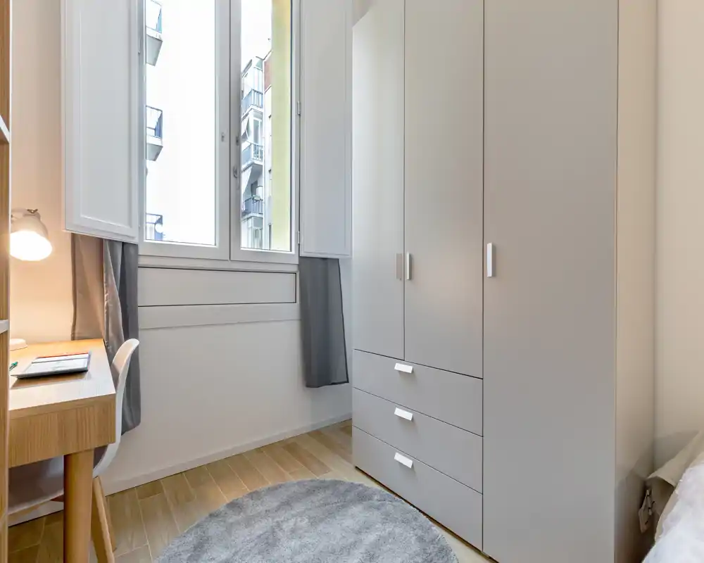 Rincón de dormitorio compacto con un gran armario, escritorio y ventana luminosa; tonos neutros y una alfombra redonda crean un área de descanso limpia y funcional.