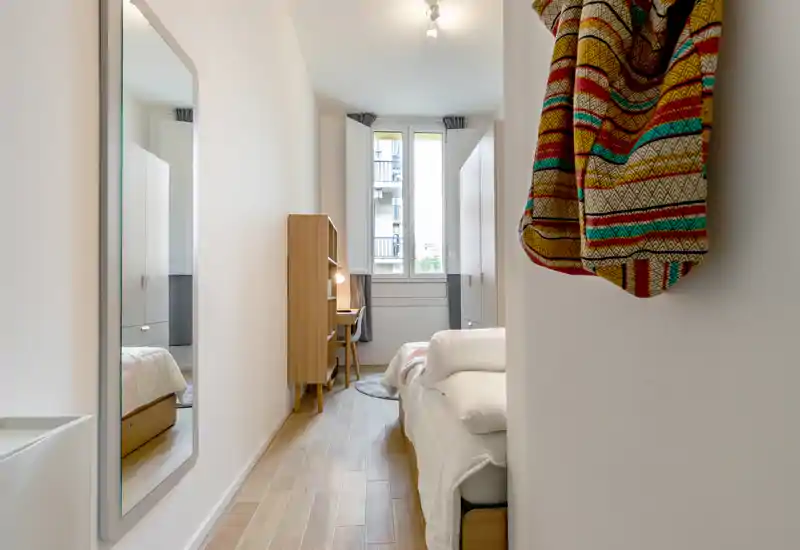 Dormitorio moderno y luminoso con cama individual a la derecha, escritorio de madera y armario junto a la ventana, y un gran espejo a la izquierda — limpio, espacioso y bien iluminado.